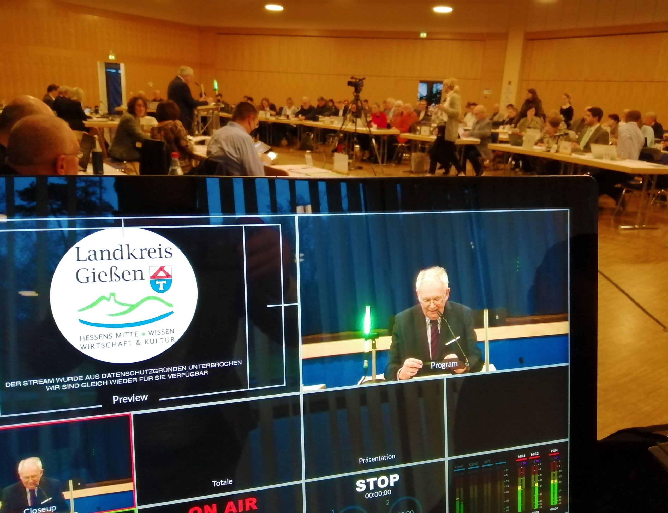 Ein Bildschirm, auf dem eine Person am Mikrofon zu sehen ist. Daneben ist das Landkreis-Gießen-Logo eingeblendet. Im Hintergrud sitzen Personen an Tischen.